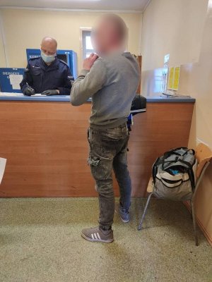 w pomieszczeniu policyjnego depozytu znajduje się umundurowany policjant i zatrzymany mężczyzna. Policjant w maseczce ochronnej na twarzy stoi za ladą i wypełnia dokumentację. Zatrzymany mężczyzna stoi po przeciwnej stronie lady