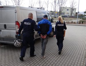 Umundurowani policjant i policjantka prowadzą zatrzymanego mężczyznę, skutego kajdankami, w kierunku nieoznakowanego radiowozu