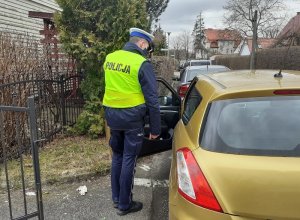 na osiedlowej ulicy przy otwartych drzwiach samochodu osobowego od strony kierowcy, stoi umundurowany policjant ruchu drogowego w kamizelce odblaskowej