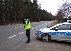 na poboczu drogi, przed radiowozem stoi umundurowany policjant ruchu drogowego w kamizelce odblaskowej. Policjant mierzy prędkość ręcznym miernikiem