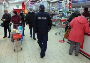 teren sklepu wielkopowierzchniowego. W kierunku kas idzie umundurowany policjant. Obok niego znajdują się klienci sklepu w maseczkach ochronnych na twarzach