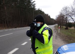 na poboczu drogi stoi umundurowany policjant ruchu drogowego w kamizelce odblaskowej i mierzy prędkość za pomocą ręcznego miernika