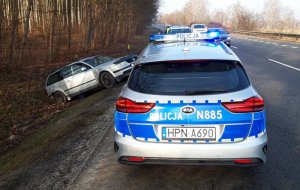 W rowie przy drodze znajduje się srebrny volkswagen passat. Na poboczu drogi, przed passatem stoi oznakowany radiowóz z włączonym sygnałami błyskowymi. W tle znajduje się policjant ruchu drogowego. Drogą przejeżdżają samochody