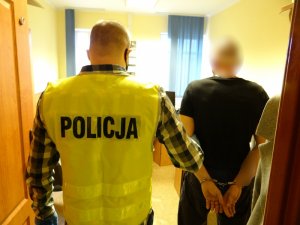 W drzwiach pokoju przesłuchań stoi zatrzymany mężczyzna z rękami skutymi z tyłu kajdankami. Po jego lewej stronie stoi nieumundurowany policjant w kamizelce z napisem policja. Policjant trzyma zatrzymanego za przedramię