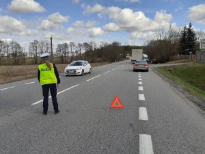 miejsce wypadku drogowego. Na pierwszym planie znajduje się policjantka, która kontroluje ruch pojazdów. Obok niej znajduje się trójkąt ostrzegawczy. W tle, na poboczu drogi widoczne pojazdy uczestniczące w wypadku: mercedes sprinter i samochód ciężarowy