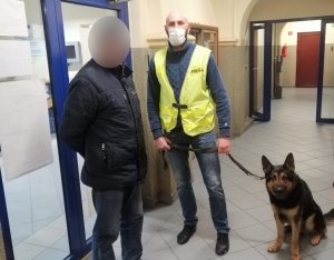 W korytarzu komendy policji stoją obok siebie odnaleziony mężczyzna i nieumundurowany policjant w kamizelce odblaskowej z napisem policja i maseczce ochronnej na twarzy. Policjant trzyma na smyczy psa służbowego