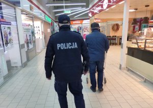 Teren sklepu wielkopowierzchniowego. Wzdłuż punktów handlowych idzie umundurowany policjant. Przed nim idzie klient pchający wózek sklepowy