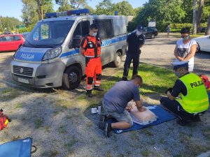 pokaz zasad resuscytacji na świeżym powietrzu. Umundurowany policjant i ratownik medyczny instruują turystę jak udzielać pierwszej pomocy medycznej. Turysta ćwiczy zasady resuscytacji na fantomie