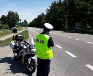 na poboczu drogi zaparkowany jest motocykl policyjny. Za nim stoi umundurowany policjant który mierzy prędkość za pomocą ręcznego miernika prędkości