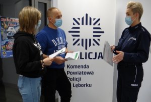 na tle roll up z logo gwiazdy policyjnej i napisem komenda powiatowa policji w Lęborku stoi umundurowana policjantka, która rozmawia z nieumundurowanymi policjantem i policjantką. Wszyscy mają maseczki ochronne na twarzach