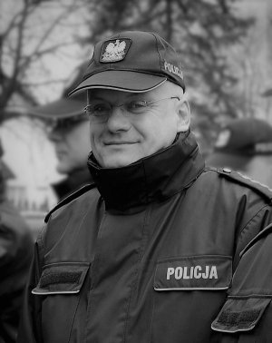 zdjęcie w czarno - białej tonacji. Na zdjęciu znajduje się uśmiechnięty policjant w stopniu aspiranta sztabowego, ubrany w służbową kurtkę i czapkę z daszkiem