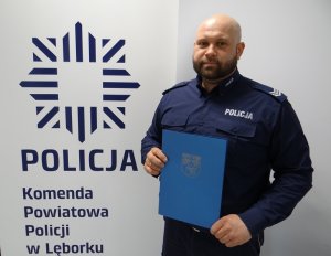 na tle logo gwiazdy policyjnej z napisem policja komenda powiatowa policji w lęborku stoi umundurowany policjant z dyplomem w rękach