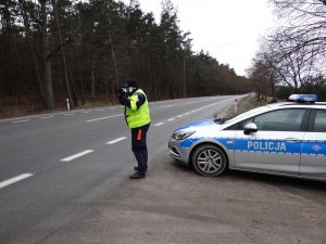 na poboczu krajowej &quot;szóstki&quot; obok oznakowanego radiowozu stoi policjant ruchu drogowego. Policjant mierzy prędkość ręcznym miernikiem prędkości