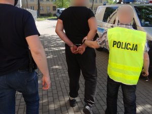 dwaj nieumundurowani policjanci, na zewnątrz budynku komendy, prowadzą zatrzymanego mężczyznę w kierunku radiowozu