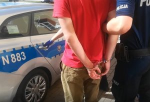 przed radiowozem umundurowany policjant trzyma za przedramię skutego kajdankami mężczyznę