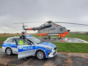 lądowisko w Łebie. Na tle wojskowego śmigłowca stoi policyjny radiowóz. Przy drzwiach radiowozu stoi umundurowana policjantka ruchu drogowego