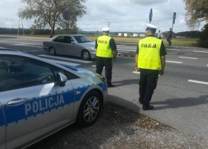 na drodze krajowej nr 6 w Leśnicach przy przejściu dla pieszych przed oznakowanym radiowozem stoją dwaj policjanci ruchu drogowego. Po drugiej stornie przejścia znajduje się dwoje dzieci