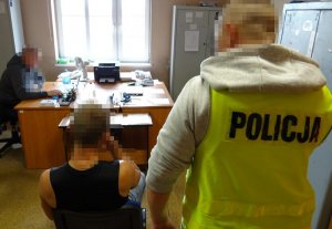 mężczyzna siedzi na krześle policjant w kamizelce stoi przy nim a drugi policjant siedzi przy biurku