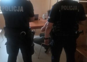 w pokoju na terenie komendy policji na krześle siedzi zatrzymany mężczyzna. Za nim stoją dwaj umundurowani policjanci