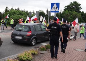 przed rondem na drodze W-214 przy wjeździe do Łeby, przez przejście dla pieszych przechodzi grupa protestujących osób z flagami i transparentami. Na przejściu dla pieszych stoi samochód. Policjant ruchu drogowego wskazuje drogę kierowcy. Po prawej stronie przejścia dla pieszych znajduje się dwóch umundurowanych policjantów oddziałów prewencji