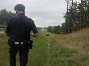 na poboczu drogi W-214 za barierami energochłonnymi stoi policjant ruchu drogowego. W odległości kilkunastu metrów przed nim, znajduje się samochód osobowy