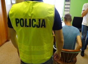 W pokoju przesłuchań na krześle siedzi zatrzymany mężczyzna. Jego ręce skute są z tyłu kajdankami. Za nim stoi policjant w kamizelce odblaskowej