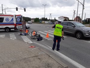 miejsce wypadku drogowego. Na przejściu dla pieszych przy skrzyżowaniu z sygnalizacją świetlną leżą dwa rowery, w tym jeden dziecięcy. To miejsce jest ogrodzone słupkami drogowymi. Przy przejściu znajduje się ambulans i policjant ruchu drogowego