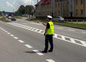 na drodze umundurowany policjant ruchu drogowego w kamizelce odblaskowej wskazuje tarczą do zatrzymywania pojazdów miejsce do zatrzymania się