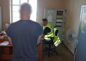 w pokoju przesłuchań, przed biurkiem przy którym siedzi policjant w kamizelce odblaskowej, stoi zatrzymany mężczyzna. Jest skierowany tyłem do obiektywu