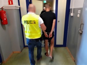 nieumundurowany policjant w kamizelce odblaskowej prowadzi skutego kajdankami mężczyznę wzdłuż korytarza komendy policji