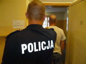 przy otwartych drzwiach do celi stoi zatrzymany mężczyzna. Jest skierowany tyłem do obiektywu. Za nim stoi policjant służby dyżurnej