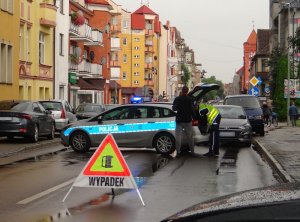 miejsce wypadku drogowego. Przejazd jezdnią jest zablokowany przez ustawiony w poprzek oznakowany radiowóz. Za radiowozem stoi osobowa toyota, za nią wóz strażacki. Do bagażnika radiowozu schyla się policjant ruchu drogowego. Przed miejscem zdarzenia na jezdni ustawiony jest odblaskowy trójkąt informujący o wypadku