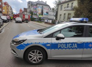 miejsce wypadku drogowego. Przejazd jezdnią jest zablokowany przez ustawiony w poprzek oznakowany radiowóz. Za radiowozem stoi osobowa toyota, za nią wóz strażacki. Policjant ruchu drogowego rozmawia z mężczyzną