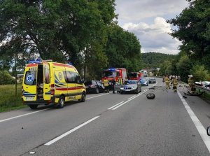 miejsce kolizji drogowej. Na poboczach po obu stronach drogi znajdują się uszkodzone samochody uczestniczące w zdarzeniu. Na miejscu jest karetka pogotowia, straż pożarna, radiowozy policyjne