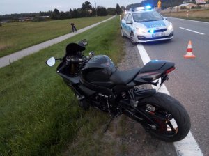 miejsce wypadku drogowego odgrodzone słupkami. Na poboczu stoi motocykl. Za nim znajduje się oznakowany radiowóz policyjny z włączonymi sygnałami błyskowymi