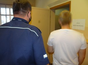 pomieszczenie dla osób zatrzymanych. Przed otwartymi drzwiami celi stoi zatrzymany mężczyzna. Za nim stoi policjant służby dyżurnej