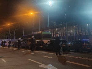 przed stadionem, wzdłuż drogi przy zaparkowanych samochodach stoją w szeregu policjanci oddziałów prewencji