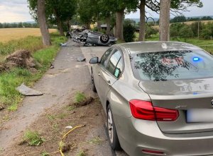 miejsce wypadku drogowego. W poprzek drogi na dachu leży audi. Auto ma widoczne liczne wgniecenia i pęknięcia karoserii, szczególnie uszkodzony jest tył pojazdu. Na drodze leżą fragmenty karoserii. Przed audi znajduje się nieoznakowany radiowóz