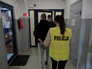 policjantka w kamizelce odblaskowej prowadzi zatrzymanego mężczyznę korytarzem na przesłuchanie. Mężczyzna ma ręce skute kajdankami