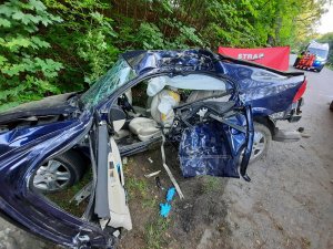 miejsce wypadku drogowego. Na pierwszym planie znajduje się rozbity o drzewo samochód, za nim rozciąga się parawan straży pożarnej, dalej na drodze stoi karetka pogotowia. Przy niej znajdują się dwaj strażacy i policjant ruchu drogowego