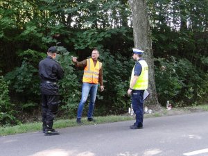 na drodze stoją policjant ruchu drogowego oraz przedstawiciel straży pożarnej i zarządu dróg wojewódzkich prowadząc rozmowę