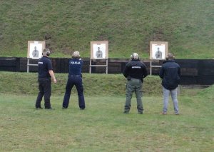 dwoje  umundurowanych policjantów oddaje strzały w kierunku tarcz. Obok nich stoją instruktorzy