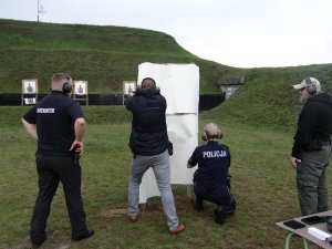 dwoje policjantów oddaje strzały zza przeszkody. Policjantka jest w pozycji klęczącej, znajdujący się po jej lewej stronie policjant wychyla się zza przeszkody z bronią skierowaną w tarczę. Za nimi stoją instruktorzy