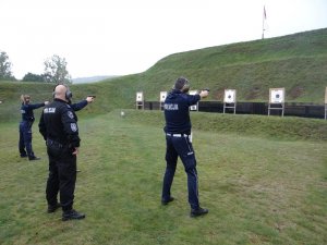 ustawieni w szeregu policjanci oddają strzały w kierunku tarcz. Za nimi stoi instruktor