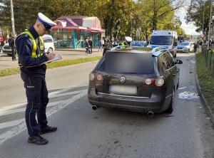 miejsce wypadku drogowego. Policjant ruchu drogowego stoi przy uczestniczącym w wypadku volkswagenie, wypełniając dokumentację. Drug policjant dokonuje oględzin pojazdu. Za nimi stoi oznakowany radiowóz