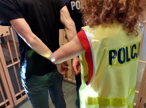 w korytarzu pomieszczenia dla osób zatrzymanych policjantka w kamizelce z napisem policja zakłada kajdanki zatrzymanemu mężczyźnie