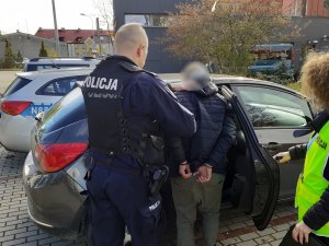 policjant wprowadza do nieoznakowanego radiowozu skutego kajdankami mężczyznę. Drzwi auta przytrzymuje nieumundurowana policjantka w kamizelce z napisem policja