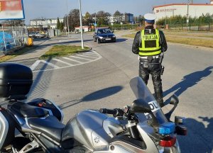 policjant ruchu drogowego stoi na drodze dojazdowej do cmentarza kontrolując ruch. Za nim stoi zaparkowany motocykl policyjny