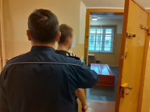 przed otwartymi drzwiami celi stoi zatrzymany mężczyzna. Za nim stoi policjant służby dyżurnej