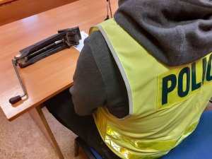 Przy stole siedzi nieumundurowany policjant w kamizelce z napisem policja. Przed nim na stole leży lewarek samochodowy